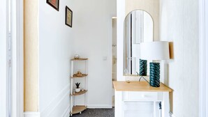 A hallway with a white wall, a wooden console table with a mirror and a lamp, and a shelving unit with decorative items.