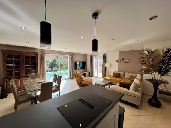 A modern living room with a dining area, a flat-screen TV, and a large window overlooking a pool.