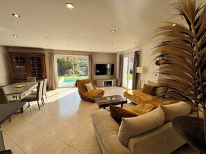 A spacious living room with a large TV, comfortable seating, a dining area, and a view of a swimming pool.