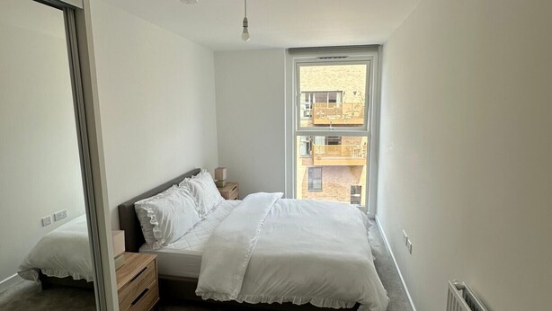A bedroom with a bed, a nightstand, a mirror, and a window with a view of buildings. - London Bliss Apartment Retreat (London)