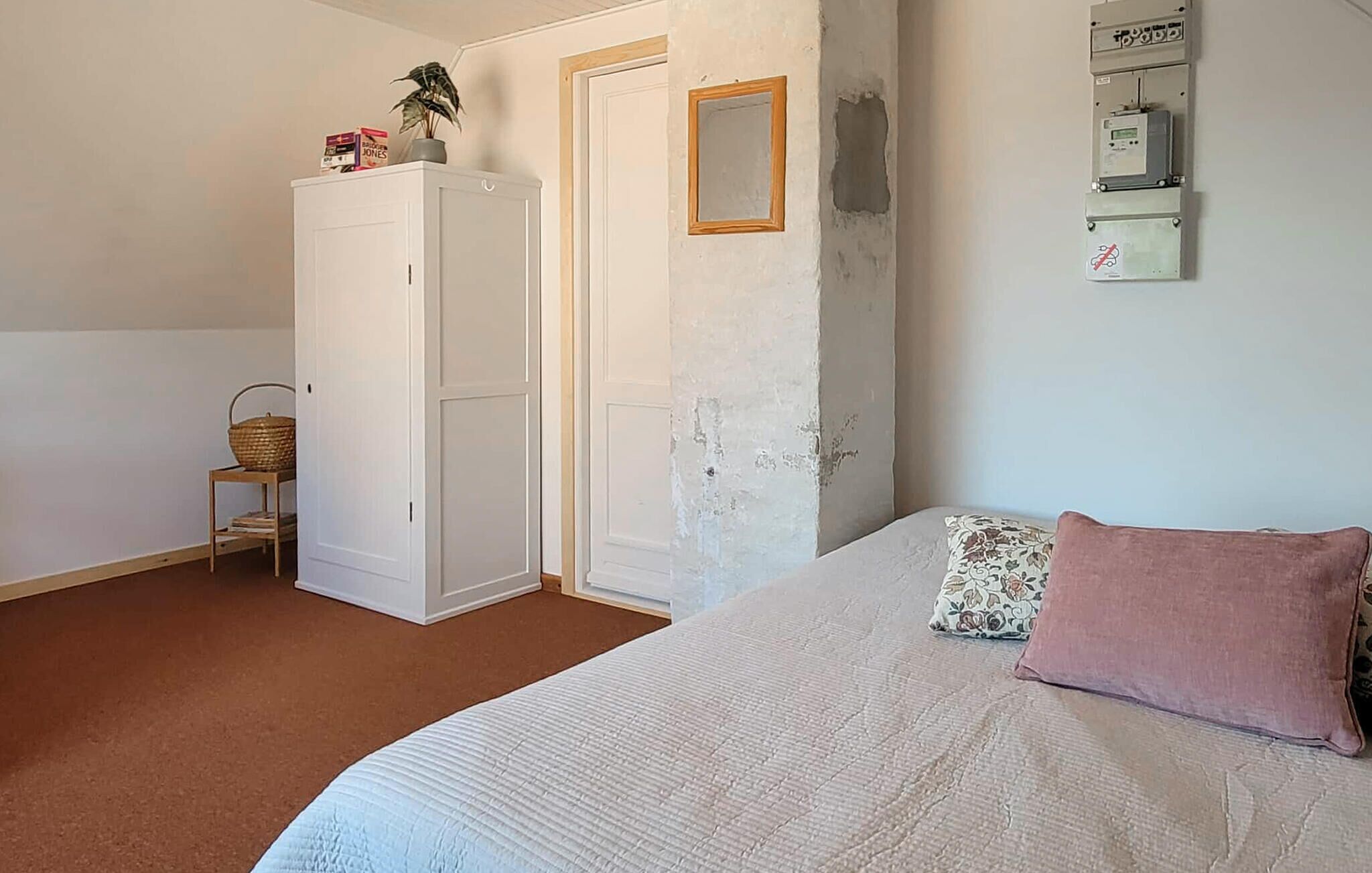 Une chambre à coucher comprenant un lit, une armoire, une petite table avec un panier, et un tableau électrique fixé au mur.