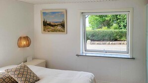 Un dormitorio con una cama, dos almohadones estampados, una lámpara de pared, un cuadro enmarcado y una ventana con vistas a zonas verdes.