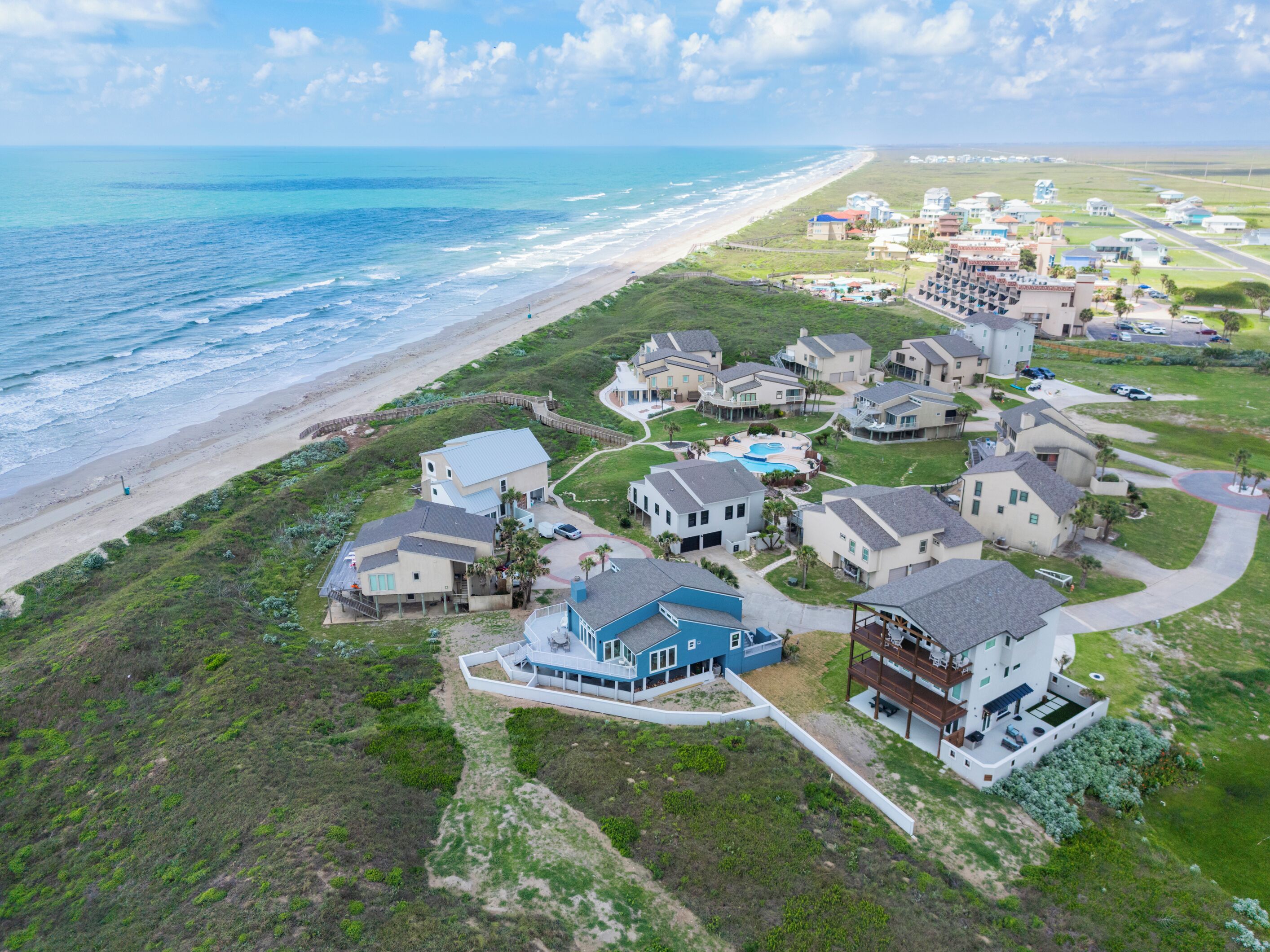 An aerial view of a coastal residential area with houses, a beach, and a pool.