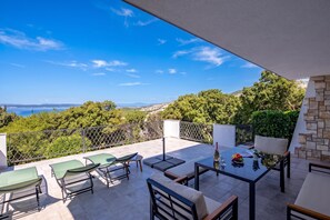 A terrace with outdoor furniture, a table with a bottle and fruit, and a view of the sea and greenery. - Hidden Pool House With Sea View (Novi Vinodolski)