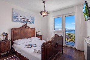 A bedroom with a large bed, a painting, a chandelier, a TV, and a balcony with a view. - Hidden Pool House With Sea View (Novi Vinodolski)