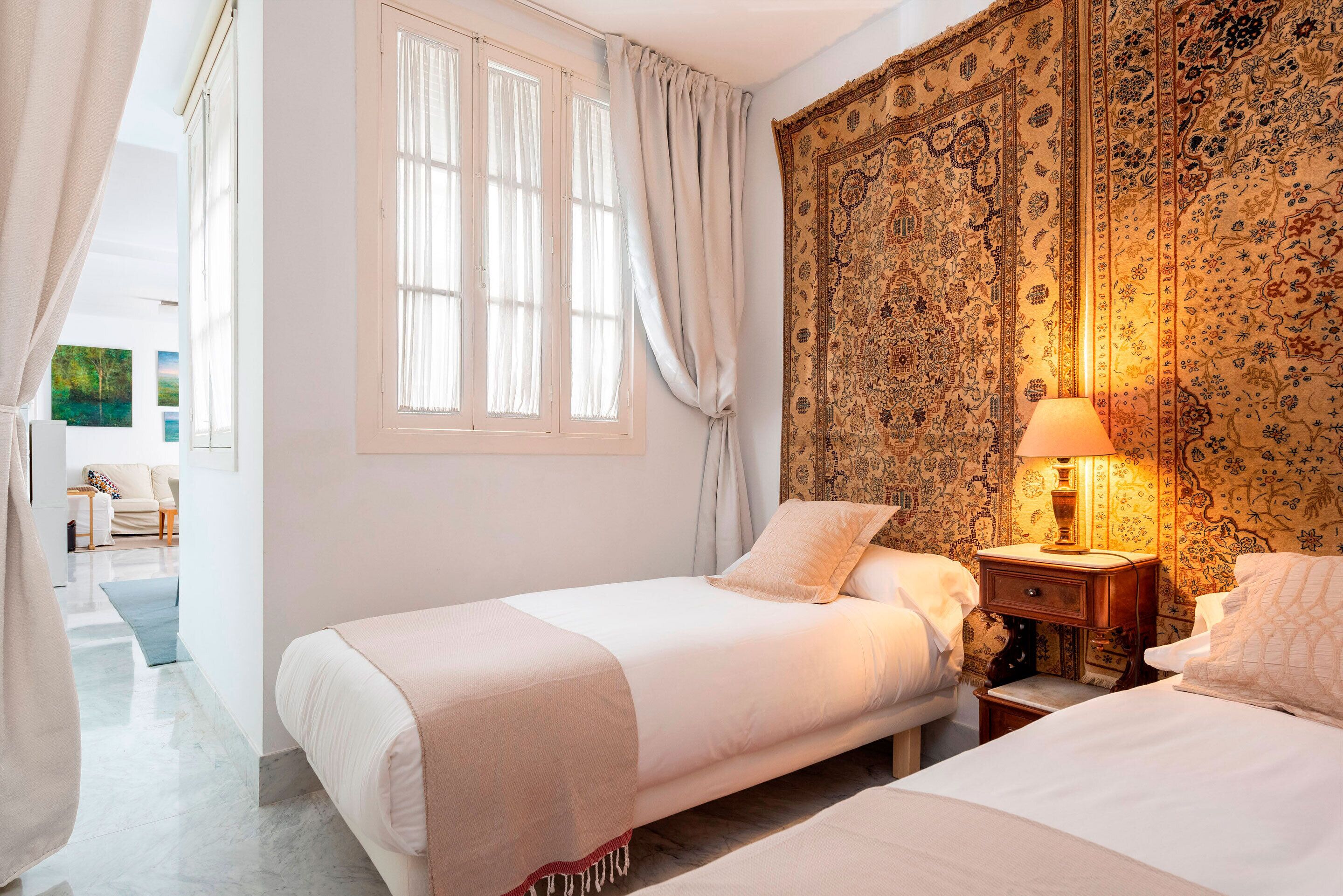 A bedroom with two beds, a window with curtains, a patterned tapestry, and a bedside lamp.