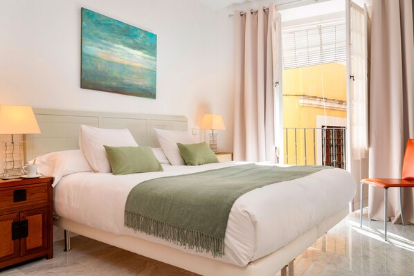 A neatly made bed with a green throw, two bedside tables with lamps, a painting on the wall, and a balcony with curtains.