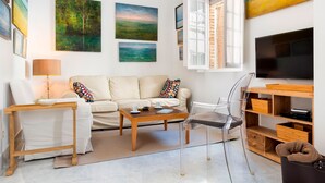 A modern living room with a sofa, coffee table, and clear chairs. There is a TV on the wall and artwork above the sofa. - Cozy Apartment in the Center. Pasaje Santa Cruz II (Seville)