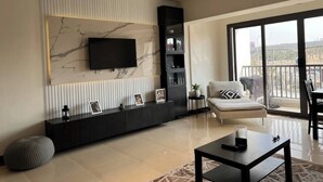A modern living room with a large TV mounted on a marble wall, a black entertainment unit, a sofa with a patterned pillow, and a view of a cityscape through large windows. - MISK properties 2 bedroom duplex 2 (Cairo Governorate)