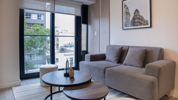 A modern living room with a grey sofa, a round wooden coffee table, and a large window offering a view of buildings and trees. - w Bright 1BR w Balcony in Miraflores (Lima)
