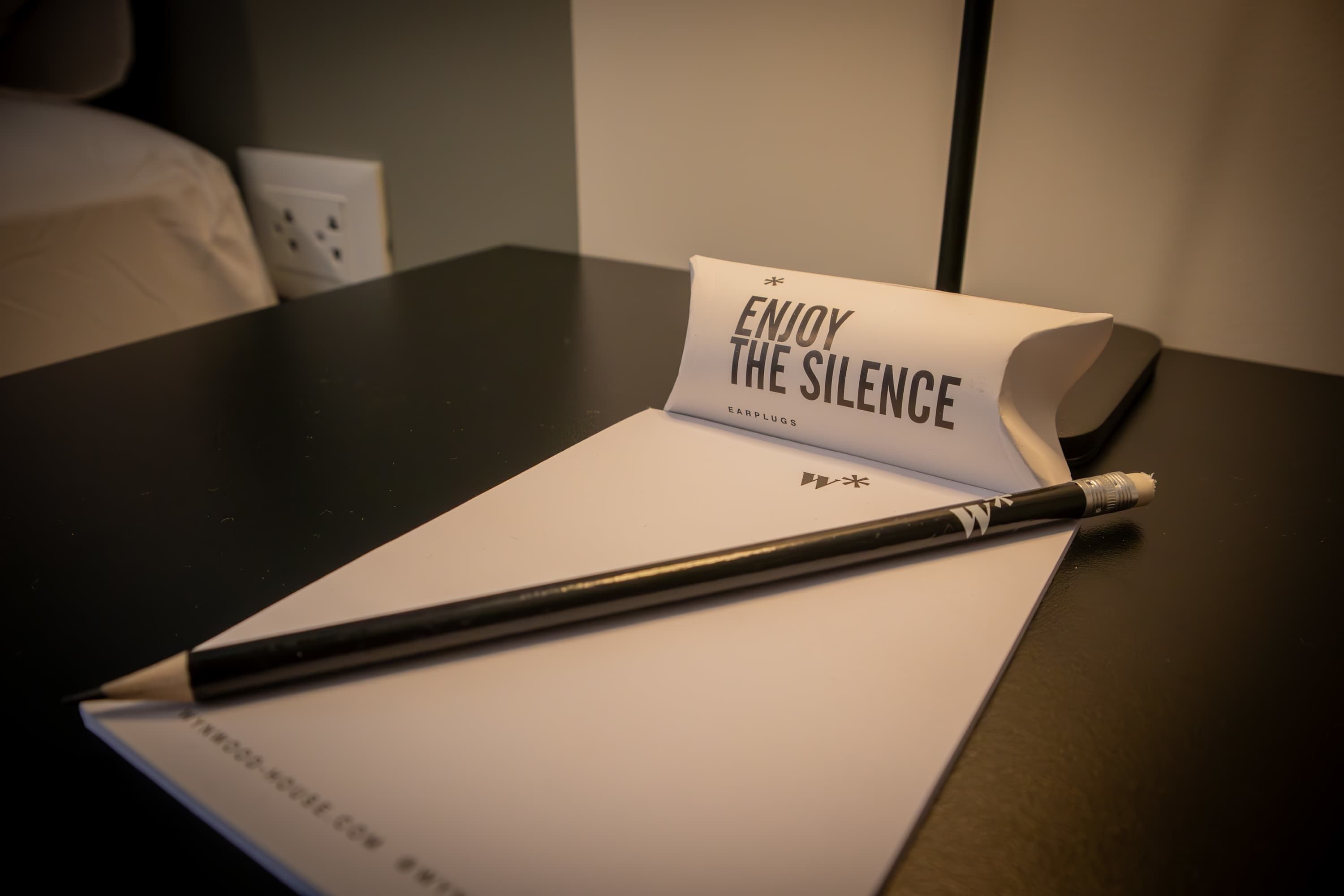 A black table with a white notepad, a black pen, and a rolled-up paper with the text "ENJOY THE SILENCE" and "EARPLUGS" on it.