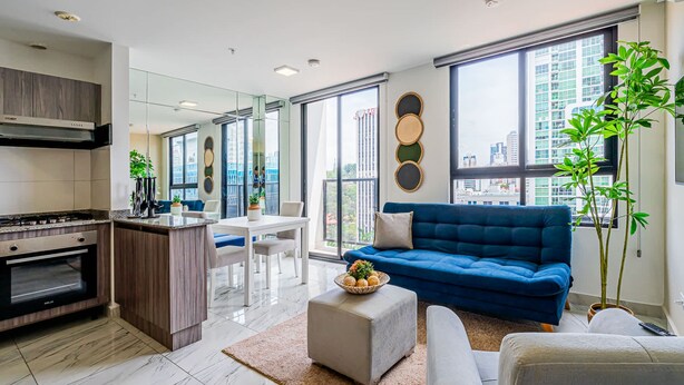Una cocina moderna con horno empotrado, una zona de comedor con mesa y sillas blancas, un sofá azul y grandes ventanales con vistas a la ciudad.