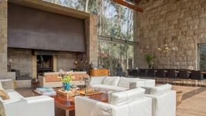 A spacious living area with a stone fireplace, white sofas, a chess table, and a view of the forest through large windows. - w Elegant 5BR Home w Pool in Cieneguilla (Pachacámac)