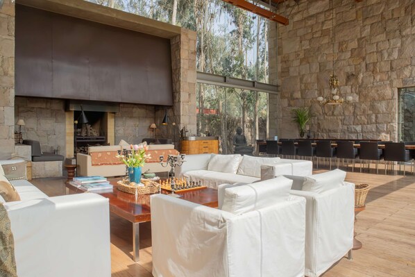 A spacious living area with a stone fireplace, white sofas, a chess table, and a view of the forest through large windows. - w Elegant 5BR Home w Pool in Cieneguilla (Pachacámac)