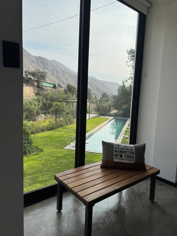 Une pièce dotée d’une grande fenêtre donnant sur une piscine et des montagnes. Un banc en bois avec un coussin se trouve au premier plan.
