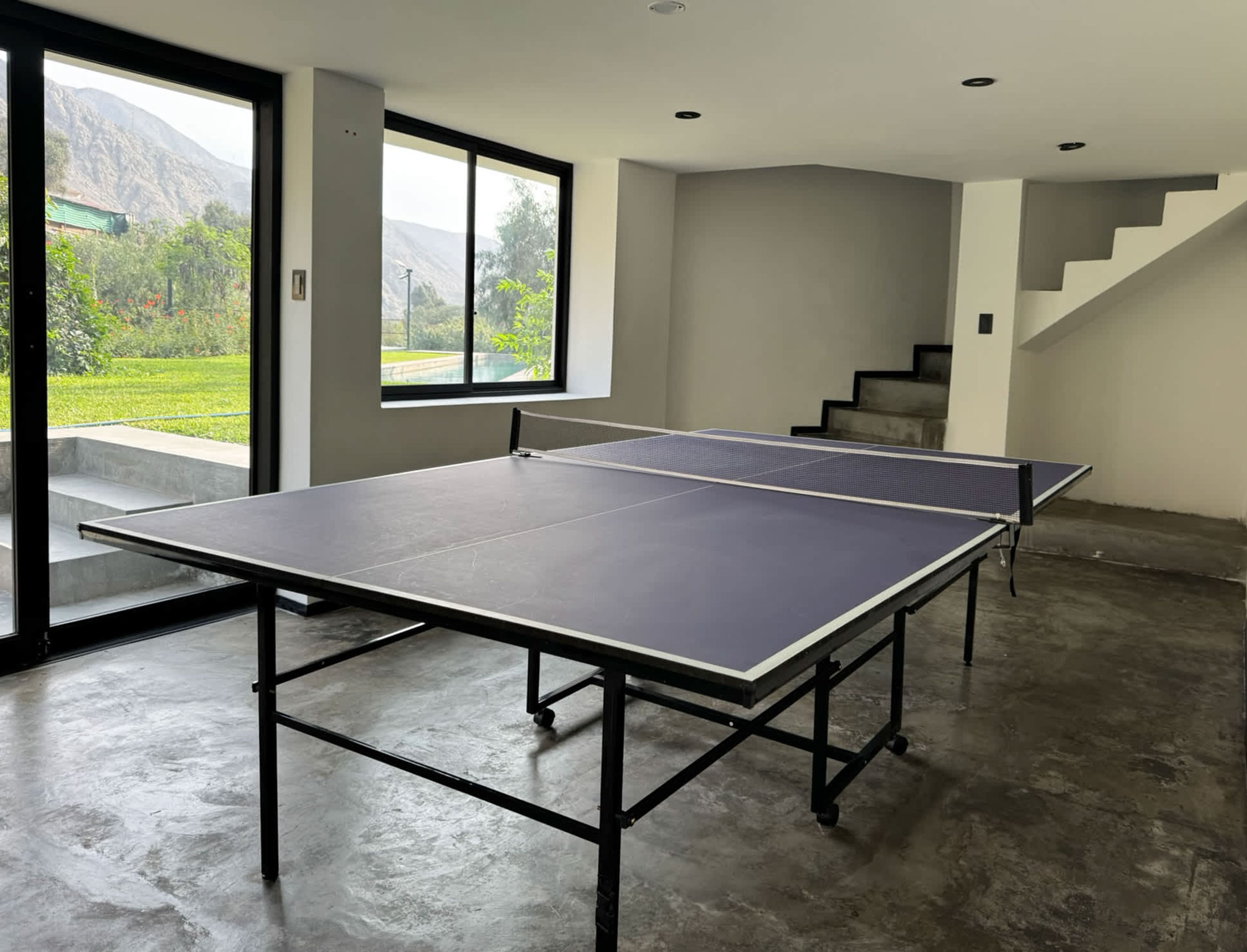 A ping pong table in a room with a large window, a staircase, and a concrete floor.