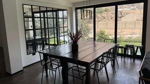 Un comedor con una larga mesa de madera, sillas negras y un jarrón de plantas secas. Amplias ventanas ofrecen vistas a un jardín ajardinado y un muro de piedra.