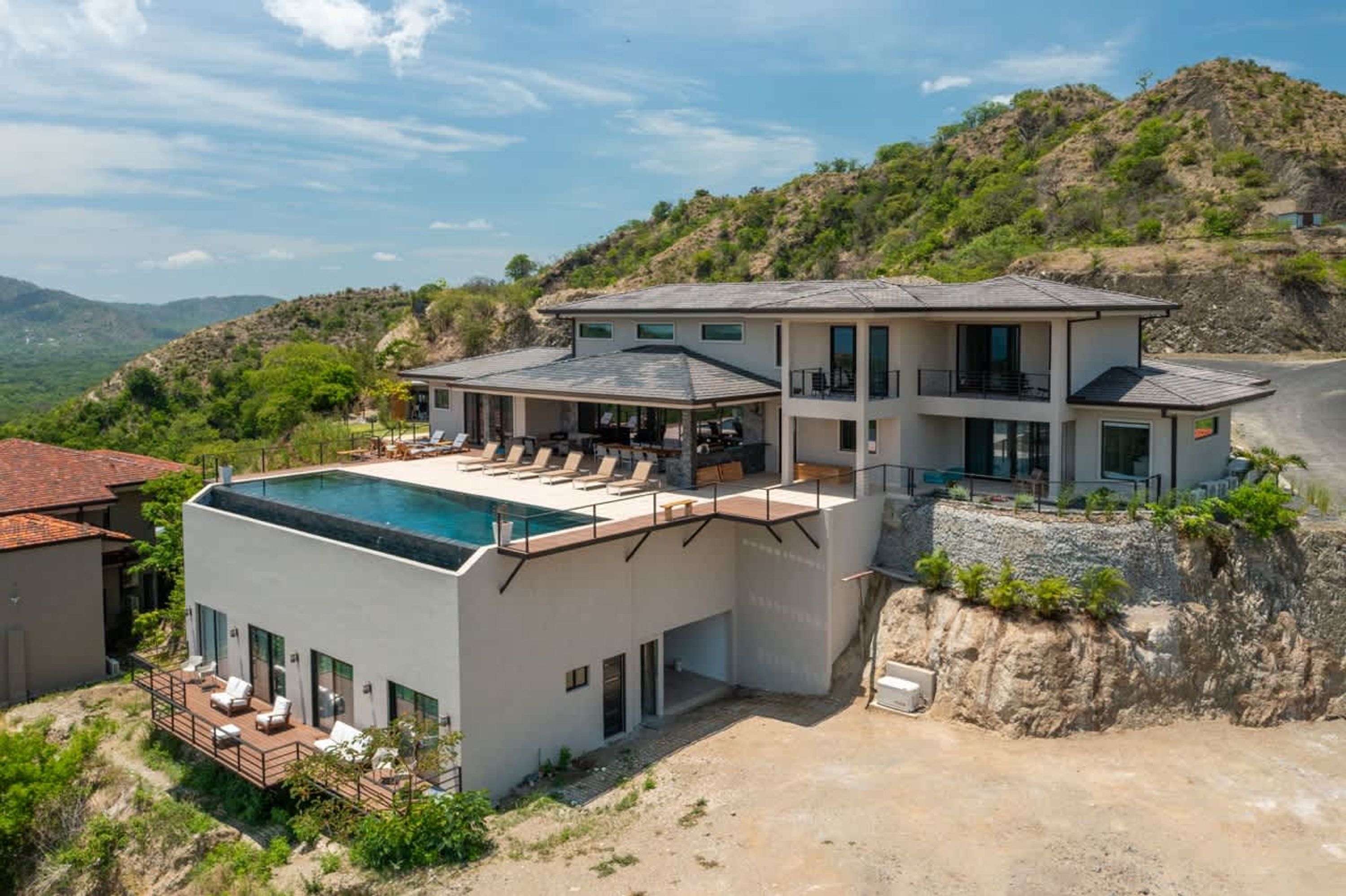 A modern two-story house with a rooftop pool, surrounded by hills and a clear sky.