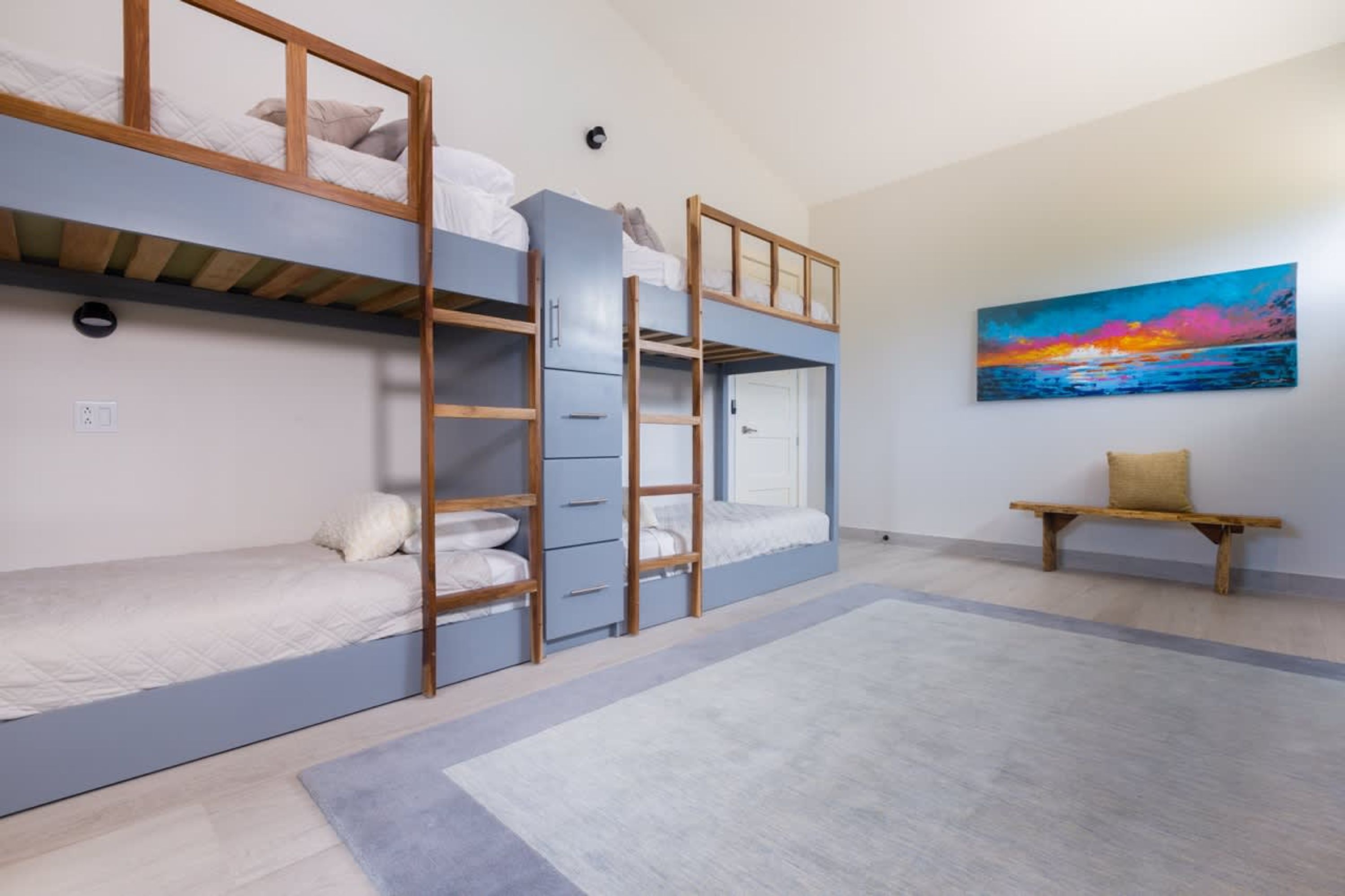Une chambre de dortoir avec des lits superposés, un tableau représentant un coucher de soleil et un banc.