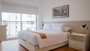 A modern hotel room with a large bed, a bedside table with a lamp, a framed cityscape picture on the wall, and a view of the city through the window. - Stunning 1BR With Great Deco in Miraflores (Lima)