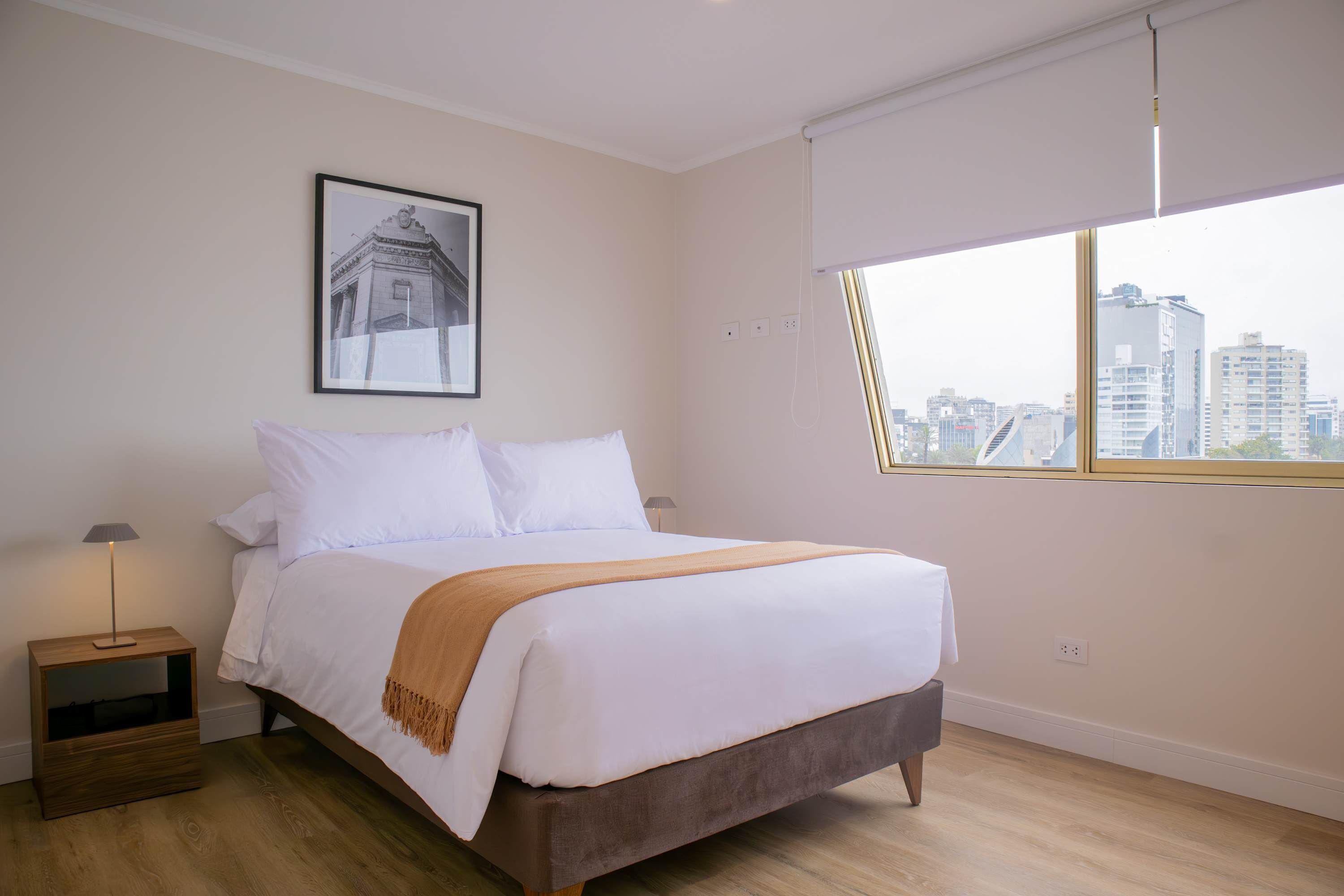 A bedroom with a bed, a nightstand, a lamp, a window with a view of buildings, and a framed picture on the wall.