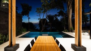 An outdoor pool area with a wooden table and chairs, surrounded by trees and a view of the ocean at dusk. - w Spectacular 4BR Villa in Costa Rica (Jacó)