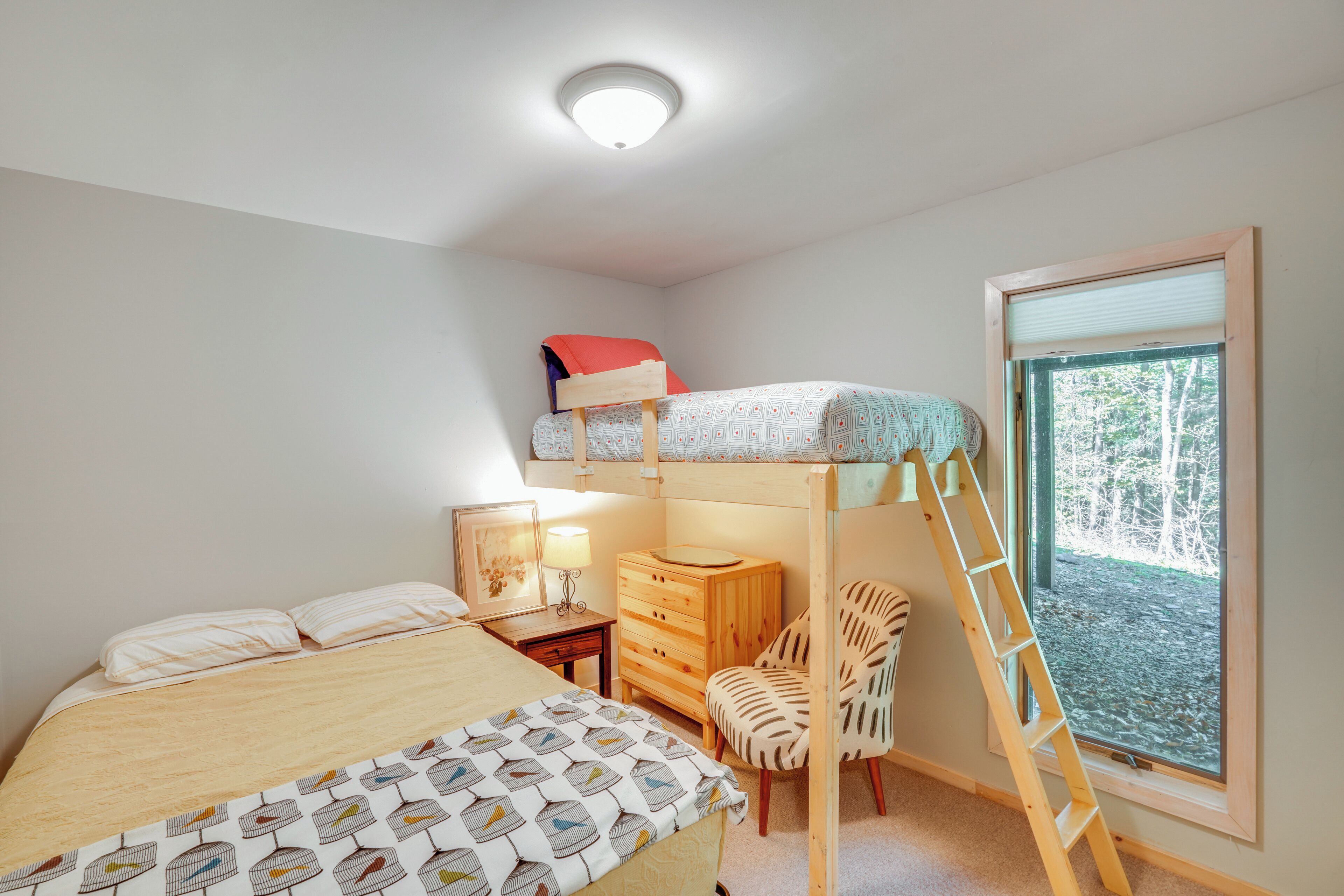 Un lit superposé avec une échelle, un bureau avec une lampe, une chaise et une fenêtre donnant sur les arbres.