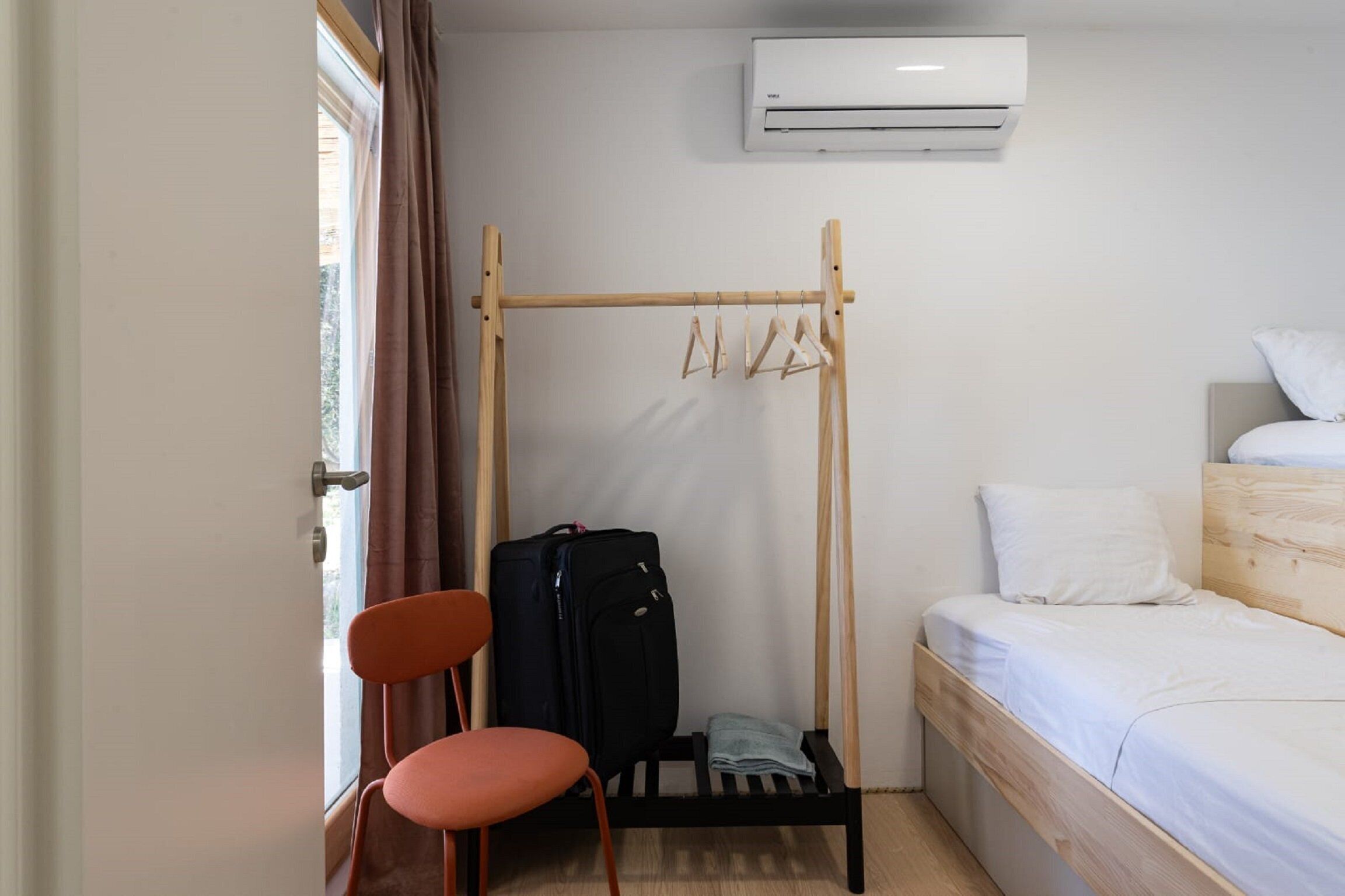 A small, tidy room with a single bed, a wooden chair, a hanging clothes rack, and a suitcase on a small table.