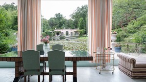 A dining area with a glass table, chairs, and a view of a garden and house through curtains. - Tranquil 1BR Villa in Revislate (Veruno)