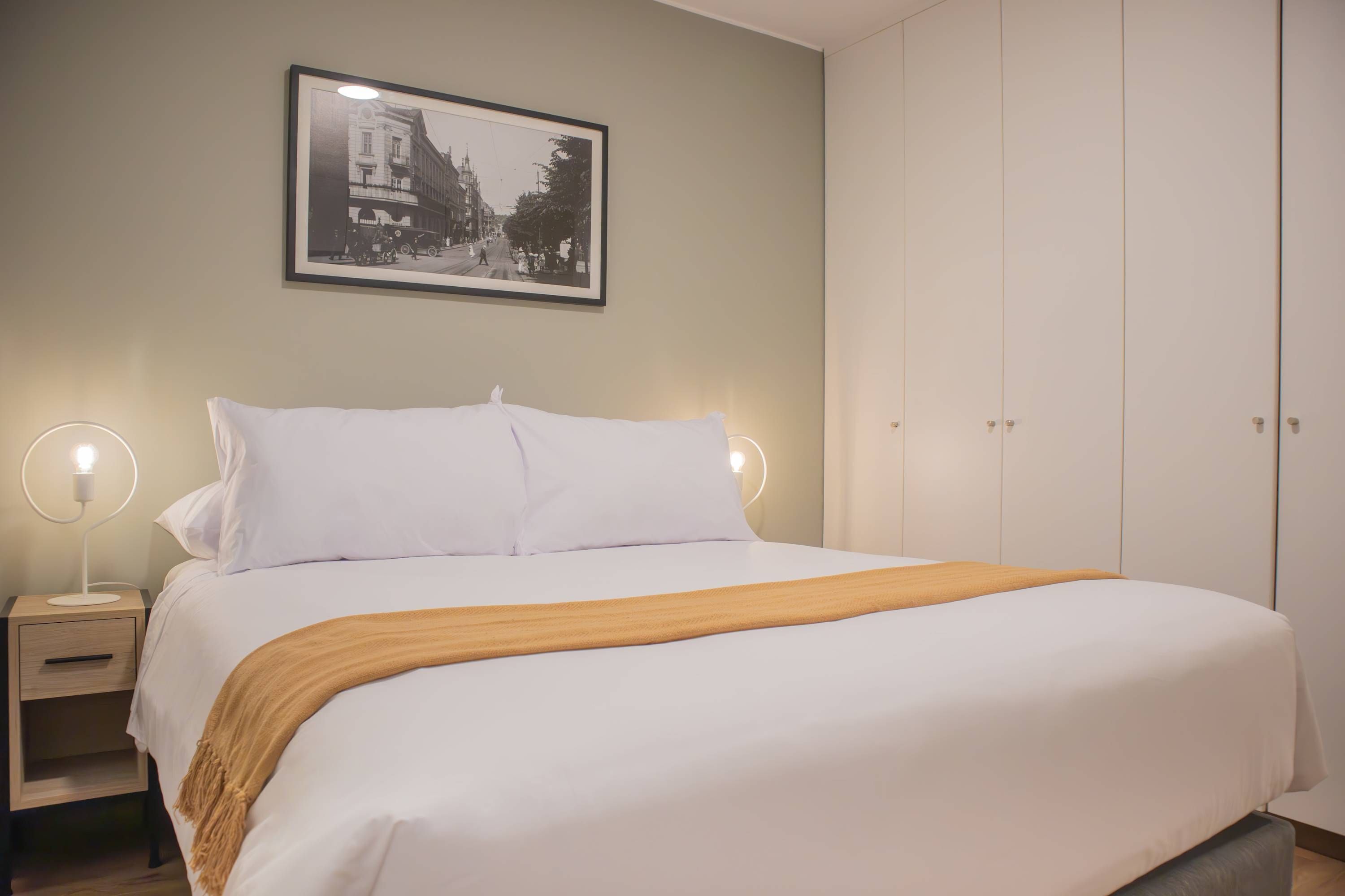 Ein ordentlich bezogenes Bett mit weißen Leinen und einer braunen Tagesdecke, ein Nachttisch mit Lampe sowie ein gerahmtes Schwarz-Weiß-Foto an der Wand.