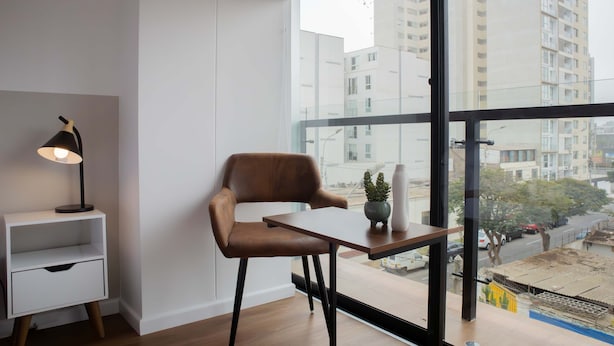 Une chambre moderne avec une grande fenêtre, un fauteuil en cuir marron, une petite table, une table de chevet blanche et un lampadaire.