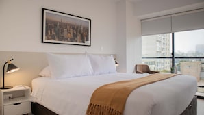 Une chambre d’hôtel avec un grand lit, une table de chevet avec une lampe, un tableau représentant un paysage urbain encadré au mur et une vue sur les bâtiments par la fenêtre.