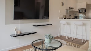 Un salon moderne doté d’un téléviseur à écran plat, d’une table basse agrémentée d’un vase, et d’un coin cuisine avec des tabourets de bar.