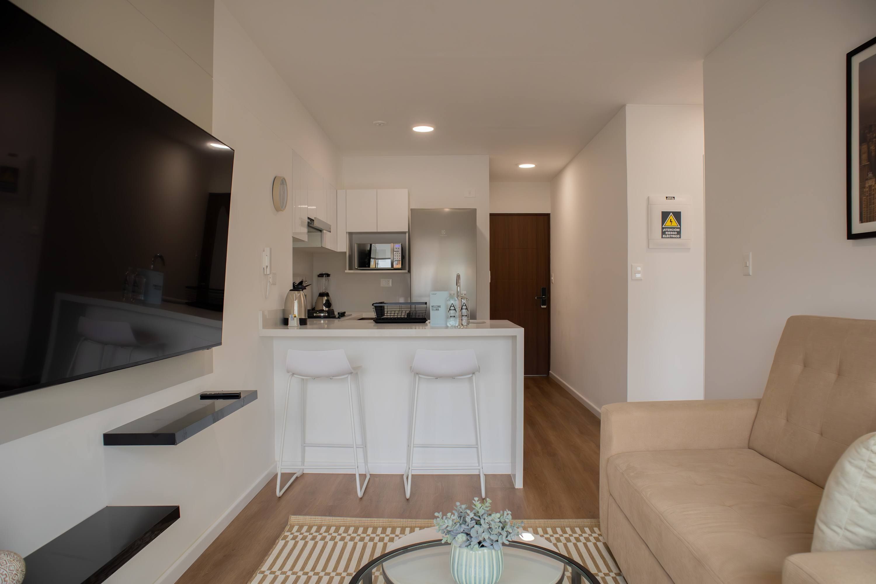 A modern living room with a flat-screen TV, a beige sofa, and a small coffee table with a vase. A kitchen with white bar stools and a microwave is visible in the background.