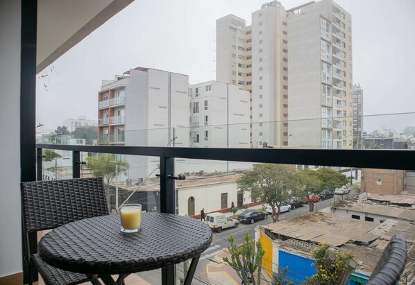 Un balcon avec une table en osier et un verre de jus d’orange, donnant sur une rue animée par la circulation des voitures et bordée d’immeubles.