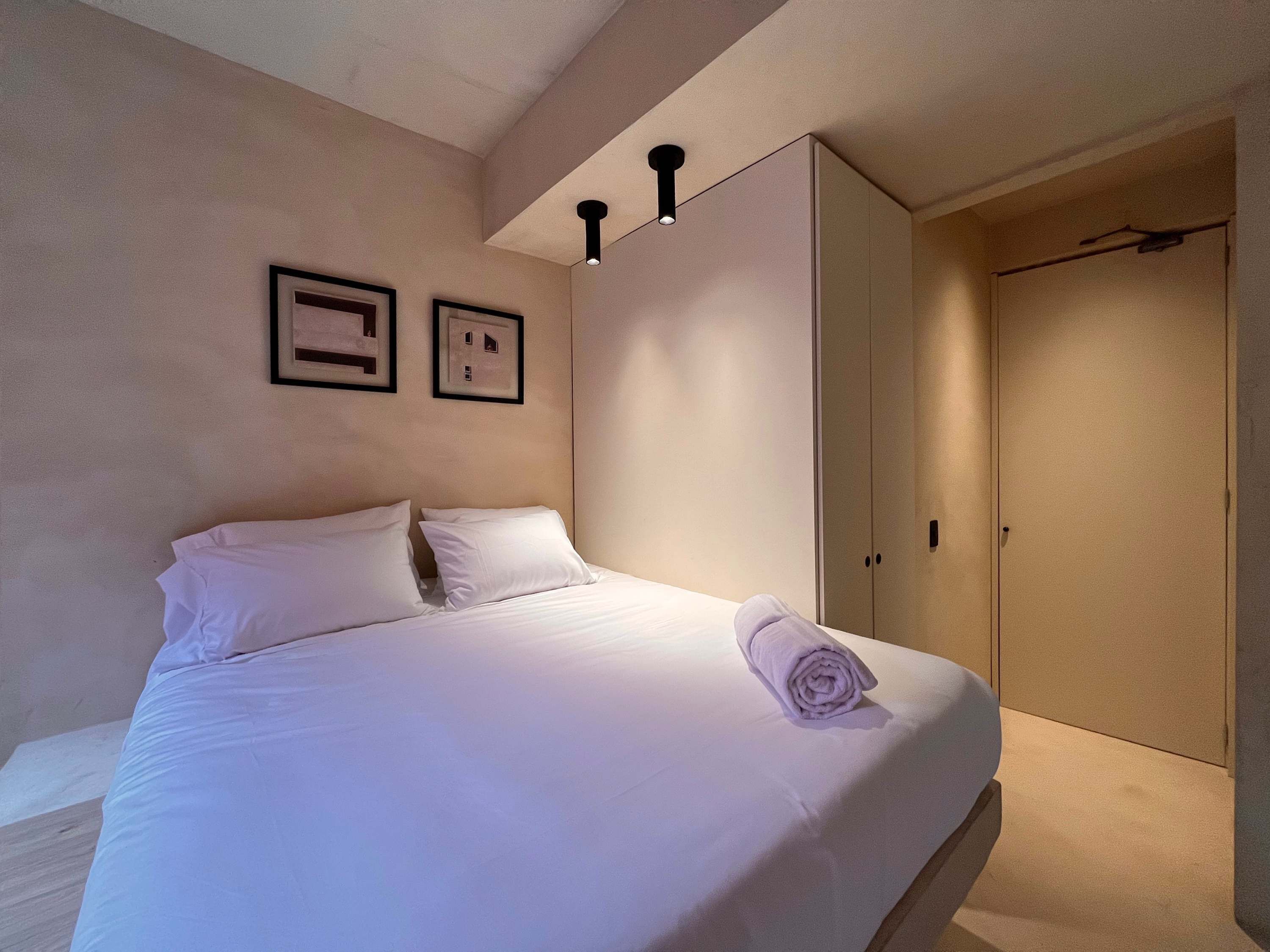 Ein ordentlich bezogenes Bett mit weißen Leinentüchern und einem aufgerollten Handtuch, ein Kleiderschrank mit Spiegel sowie zwei gerahmte Bilder an der Wand.