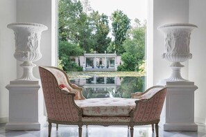 A room with a view of a garden through a large window, featuring a white columned structure and a fountain. - Charming 1BR Suite in Revislate (Veruno)