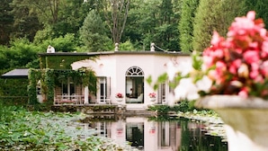 A serene pond with lily pads and a house with a white facade and arched windows, surrounded by lush greenery. - Charming 1BR Suite in Revislate (Veruno)