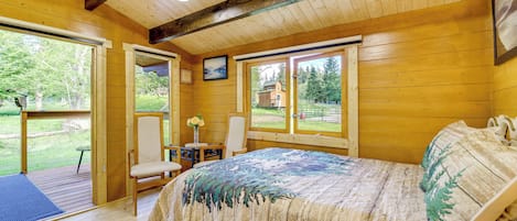 A wooden cabin bedroom with a bed, a chair, a small table, and a window overlooking a yard.