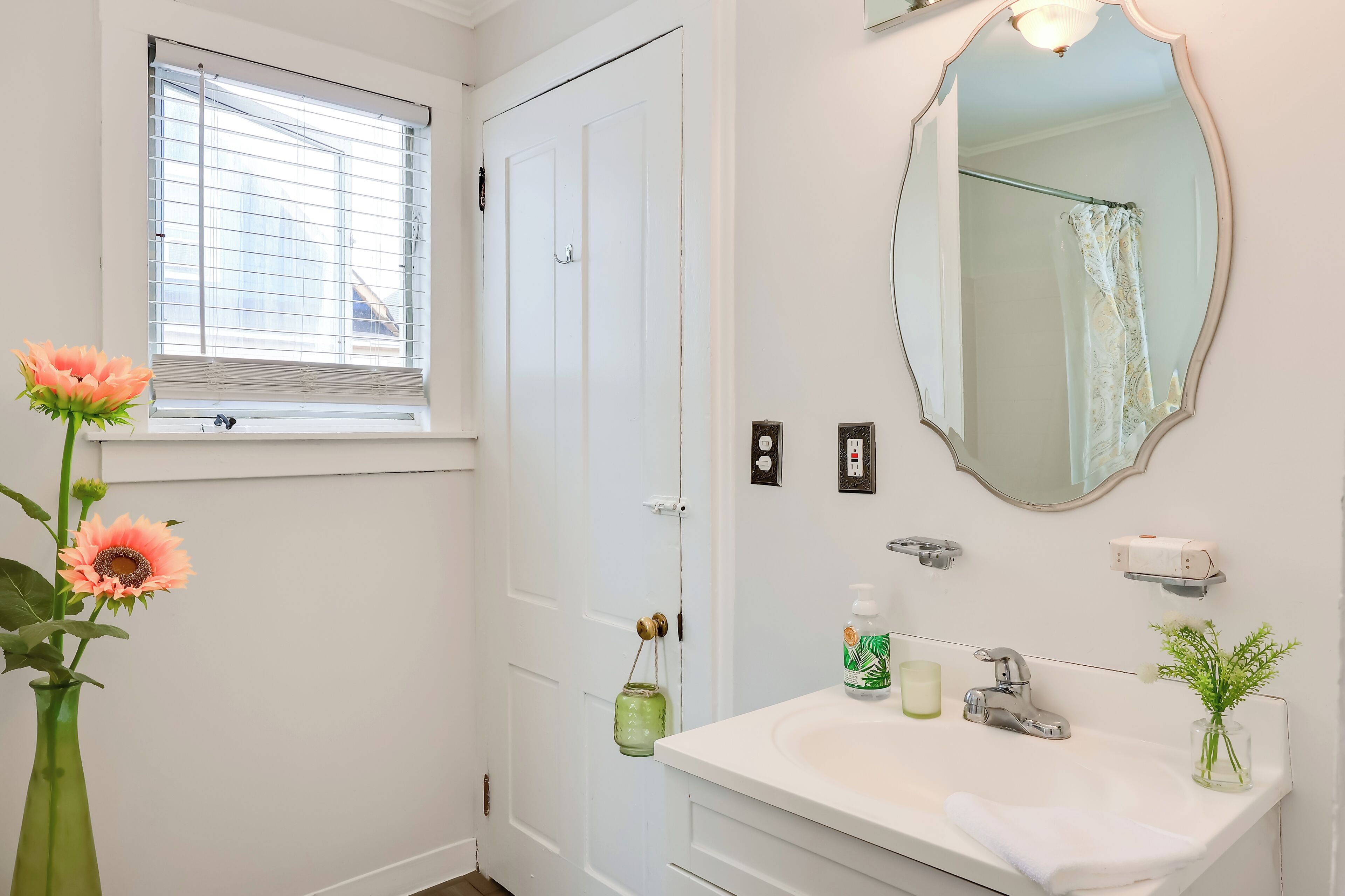 A bathroom with a white sink, a mirror, a window with blinds, and a vase with flowers.