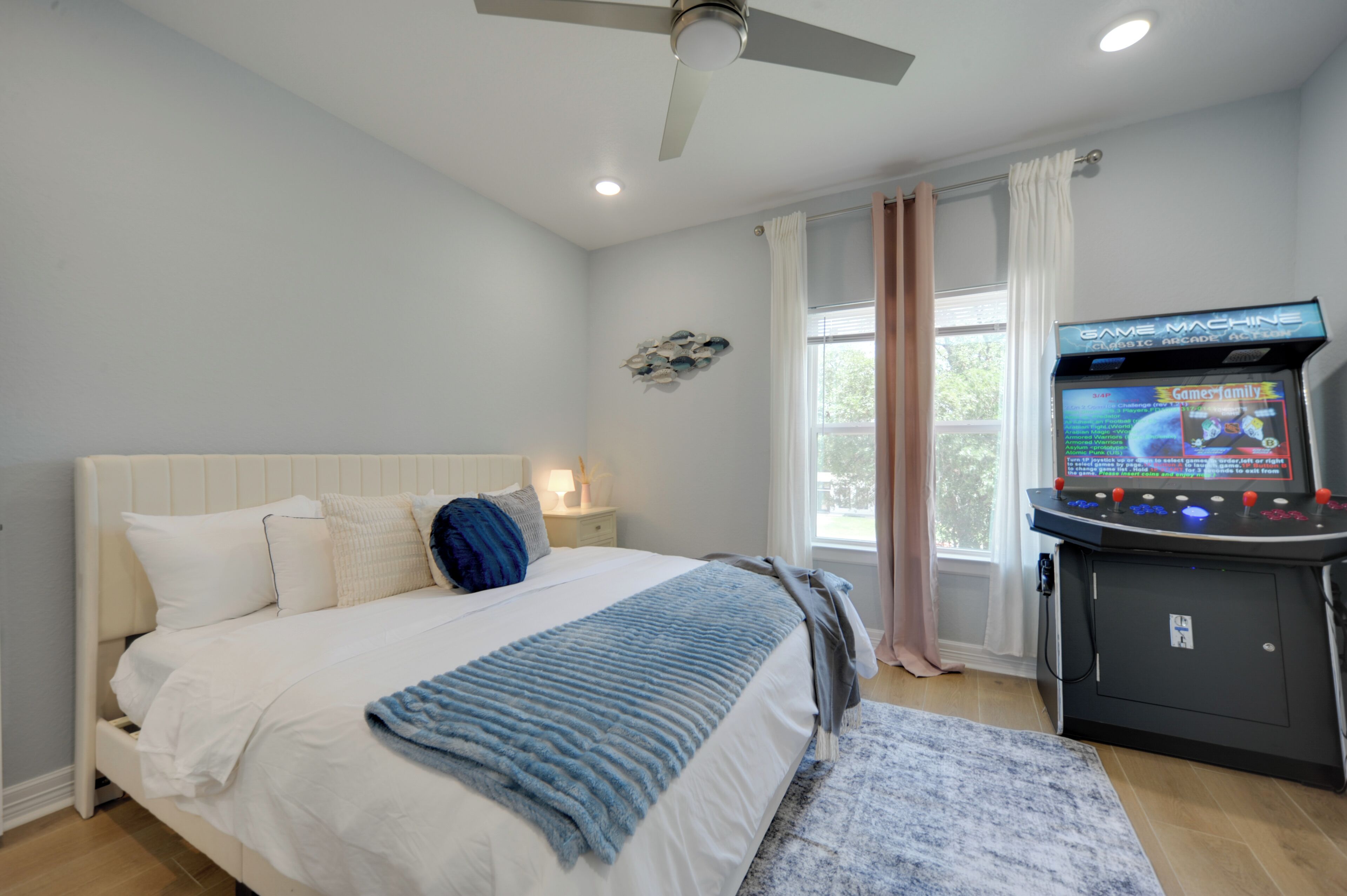A bedroom with a bed, a ceiling fan, a nightstand, a lamp, a window with curtains, and a game machine.