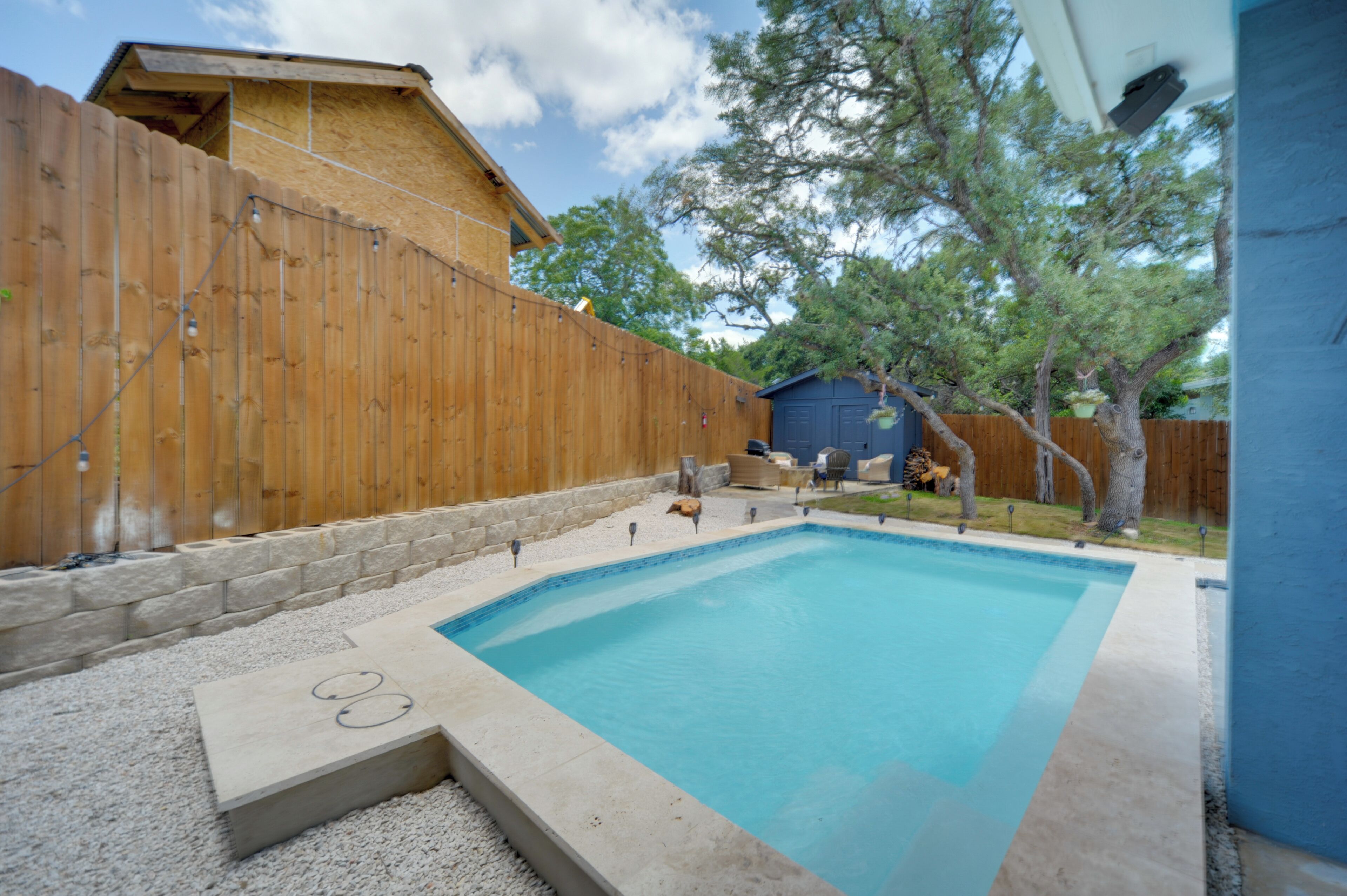 A swimming pool with a wooden deck, a stone wall, and a blue house in the background.