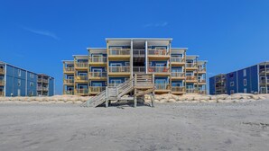 A multi-story beachfront building with balconies, a staircase leading to the entrance, and a sandy foreground.