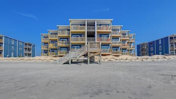 Een meerhoog gebouw aan het strand met balkons, een trap die naar de ingang leidt en een zandig voorplan.