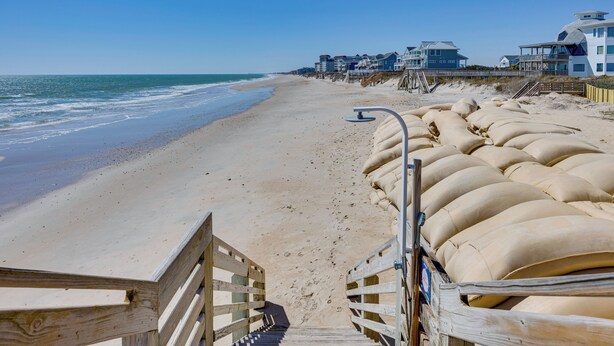 A beach with a wooden walkway leading to a sandy area. There are houses in the background.