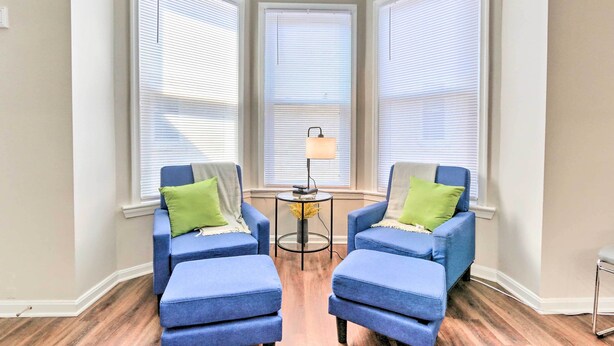 A corner room with two blue armchairs and ottomans, a small table with a lamp, and three large windows with blinds. - Central Front Royal Getaway on Main Street! (Front Royal)