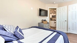 A neatly made bed with a striped bedspread, a television mounted on the wall, a kitchen area visible through an open door, and a gray nightstand with a basket. - Central Front Royal Getaway on Main Street! (Front Royal)