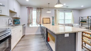 Eine moderne Küche mit weißen Schränken, einer Granit Arbeitsplatte und einem eingebauten Backofen. Zwei Pendelleuchten, ein Fenster mit Vorhängen und eine kleine Kücheninsel mit Spüle sind vorhanden.