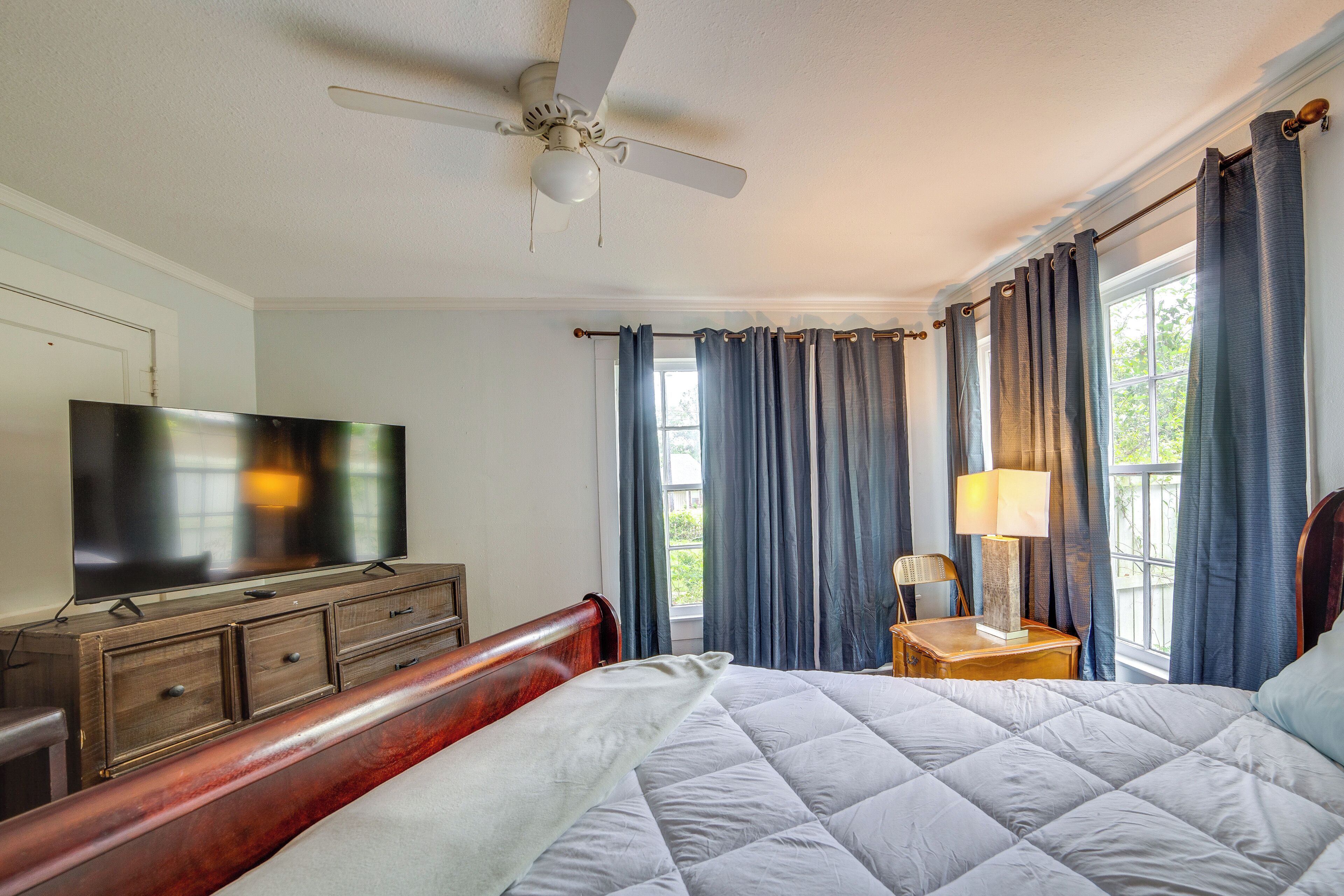 A bedroom with a bed, a TV, a dresser, a ceiling fan, and curtains.