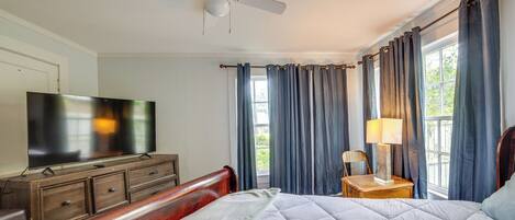 A bedroom with a bed, a TV, a dresser, a ceiling fan, and curtains.